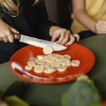 Two children preparing sliced bananas on a red plate in a kitchen setting.